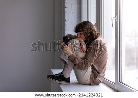 Curly young woman with a phone in her hands sits on the windowsill.