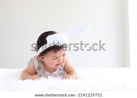 Portrait of a six months crawling baby on fluffy white rug, adorable sweet little girl kid with rabbit ears headband crying on bed in bedroom, childhood and Easter decoration concept 