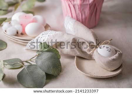 In an Easter napkin in the form of rabbit ears, a white egg with a pattern of a black branch, on a light plate.  Rustic decor.  A sprig of eucalyptus, white eggs on a linen tablecloth, pink elements.
