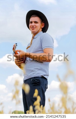 Defocus Hipster musician. Young caucasian man playing ukulele outside. Handsome hipster guy plays ukulele guitar nature background. Male in a cowboy hat smiles and plays guitar. Out of focus.