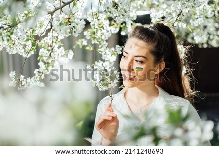 Outdoor portrait of ukrainian woman with blue and yellow ukrainian flag on her cheek on cherry blossom nature. Symbol of freedom and independence of Ukraine. Stop the war. Hope and Faith