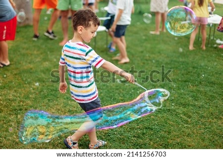 the child inflates large soap bubbles. Street animation program, happy child on a green lawn. Summer vacation. A boy in bright striped clothes with soap bubbles. Large portrait, horizontal