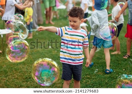 the child inflates large soap bubbles. Street animation program, happy child on a green lawn. Summer vacation. A boy in bright striped clothes with soap bubbles. Large portrait, horizontal