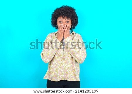 Vivacious young girl with afro hairstyle wearing floral shirt over blue background , giggles joyfully, covers mouth, has natural laughter, hears positive story or funny anecdote