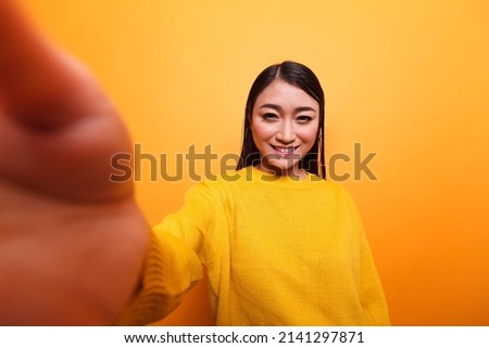 POV of happy smiling influencer wearing yellow sweater taking selfie with camera while on orange background. Pretty confident carefree vlogger smiling at camera while recording a daily vlog.