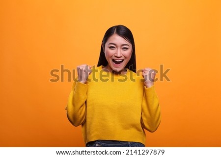 Stylish trendy happy girl celebrating victory and feeling optimistic and independent. Attractive smiling woman raising arms while feeling strong and confident on orange background. Royalty-Free Stock Photo #2141297879