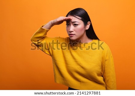 Curious intrigued asian woman holding hand above eyes and peering into distance. Young adult person with palm on forehead looking far away and searching for someone on horizon, Royalty-Free Stock Photo #2141297881