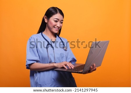 Happy smiling asian healthcare clinic nurse wearing stethoscope and using modern laptop. Young joyful caregiver staff member wearing medical instrument while using computer on orange background. Royalty-Free Stock Photo #2141297945