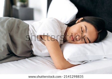 Sweet dreams. Pretty chinese woman in white t-shirt under the blanket in bed in the morning. Side view of a sleeping asian girl in a cozy bed in apartment, pleasant dreams, healthy sleep Royalty-Free Stock Photo #2141298835