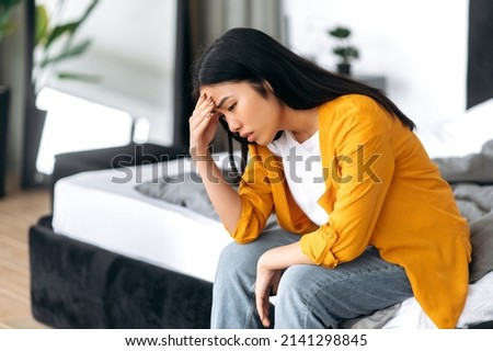 Upset sad Chinese brunette young woman, in casual clothes, sitting on the bed at home, saddened by personal problems, news, loneliness, experiencing stress, uncertainty Royalty-Free Stock Photo #2141298845