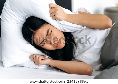 Asian young woman, lying in bed at home, covers her ears with a pillow, tries to sleep, closes herself from noise, has a bad dream, restless dream. Sleep and rest concept Royalty-Free Stock Photo #2141298861
