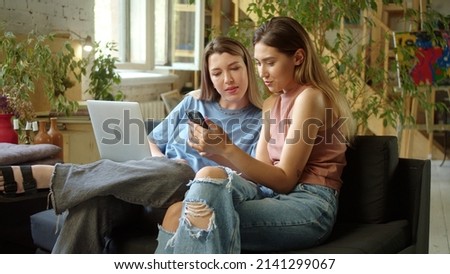 Two young beautiful ladies are searching through the Internet on the laptop, discussing and smiling