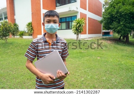 Boy student standing on grass field, children holding laptop for learinging on building school background, child wearing mask for protect viruscorona, covid-19, education concept photo for creative