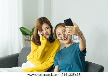 Two Pretty young asian female with big smile sitting at living room. She having fun taking light cheerful selfie on blurred background.