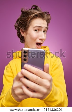 Modern teenagers. A funny emotional boy teenager in bright yellow hoodie joyfully takes a selfie. Bright purple background. Adolescents, lifestyle and communications.