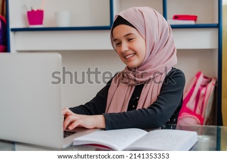 Muslim girl student in hijab smiling while learning for school at home desk using a laptop.	