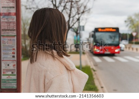 Woman from the back and standing watching her bus arrive at the bus stop. Image of a young brunette girl watching as a red bus approaches her position.