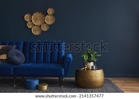 Elegant modern living room interior design with glamour blue velvet sofa, pouf, golden metal side table, plants and modern home accessories. Dark blue wall. Template. Copy space.
