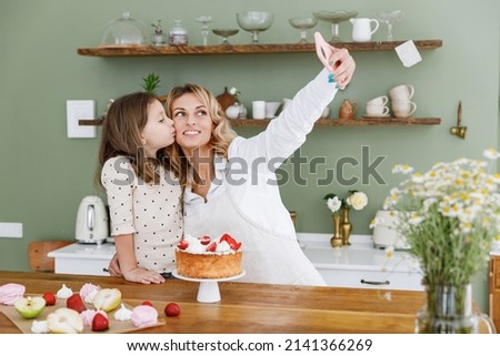 Happy chef cook baker mom woman in white shirt work baby girl helper do selfie shot on mobile cell phone kiss at kitchen table home Cooking food process concept Mommy little kid daughter prepare cake.