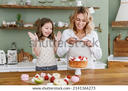 Happy chef cook baker mom woman in white shirt work with baby girl helper take photo on mobile cell phone at kitchen table home. Cooking food process concept Mommy little kid daughter prepare cake.