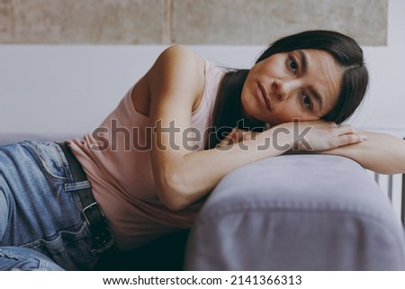 Unnerved sad worried minded young latin woman 20s in casual clothes sit lay on sofa think plan hands folded under head look camera spend time in living room home in own house. People emotions concept Royalty-Free Stock Photo #2141366313