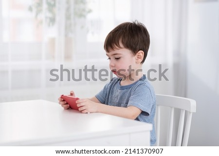 A cute little boy is sitting at the table and playing on the phone. Gadgets and kids concept