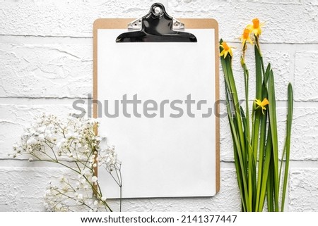 A wooden folder for documents with an iron clothespin and a white sheet of Mocap paper with spring flowers of narcissus and a sprig of gypsophila. White brick background. Royalty-Free Stock Photo #2141377447