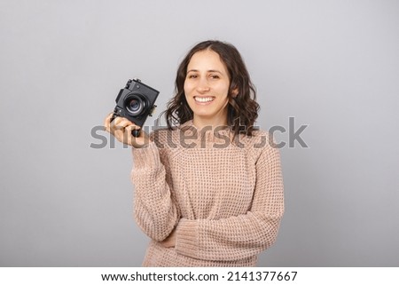 Woman photographer is holding camera in one hand while smiles at the camera. Studio shot over grey background.