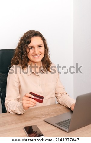 Vertical photo of a young woman holding card while working at the laptop in a white office.