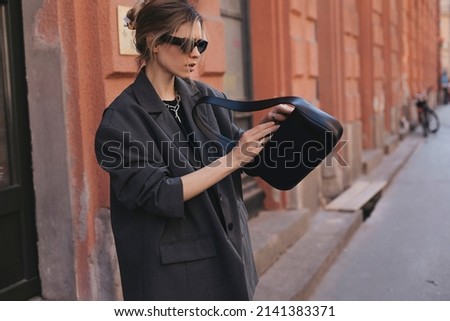 Elegant young woman looking in her black leather bag her phone or purse. Business style woman wear grey blazer, black eyeglasses and bag on the street. Street style, fashion outfit. Royalty-Free Stock Photo #2141383371