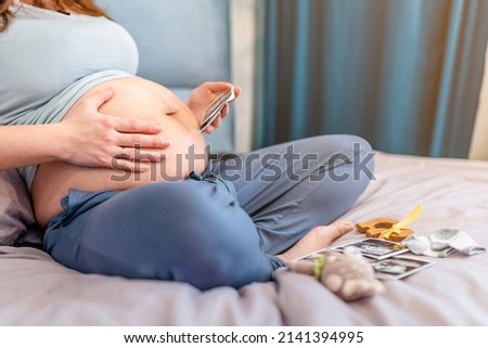 A pregnant woman is sitting on the bed and looking at ultrasound pictures of the baby, the concept of motherhood and preparation for the birth of a child