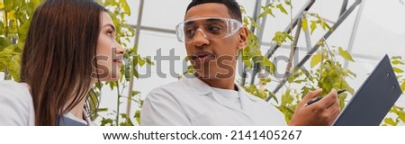 Low angle view of african american botanist holding clipboard near colleague in greenhouse, banner