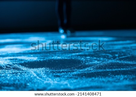 Dark silhouette of female legs in figure skating skates sliding on ice arena. Young woman trains on dark ice rink with blue light. Shiny smooth surface of ice with scratches from skates. Close up.