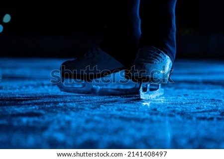 Detail shot of women legs in white figure skating skates on ice arena. Professional sportswoman trains on dark ice rink with blue light. Surface of ice with scratches and skate streaks. Close up.