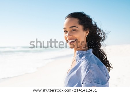 Portrait of young woman at sea looking at camera. Smiling latin hispanic girl standing at the beach with copy space and looking at camera. Happy mixed race girl in casual outfit with wind in her hair. Royalty-Free Stock Photo #2141412451