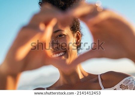 Close up of hands of young woman making heart shape. Portrait of a lovely black woman making heart with fingers while looking at camera. Smiling african american woman making love sign during sunset. Royalty-Free Stock Photo #2141412453