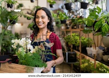 Beautiful florist holding wooden box of fresh plants at flower shop. Latin hispanic woman working in floral shop with copy space. Successful florist smiling while holding fresh flowers and plants. Royalty-Free Stock Photo #2141412459