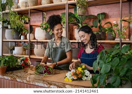 Two happy and smiling young woman florist working together in flower shop. Florist talking and making beautiful bouquet of flower together with her black colleague. Beautiful florists arranging bunch. Royalty-Free Stock Photo #2141412461