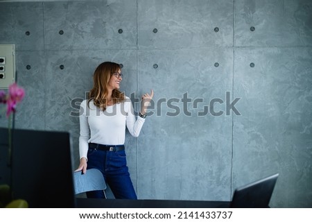 A casual, mature glasses fashion female manager is turned on her left side and pointing to a gray wall. Brainstorming and training in the corporation training hall. Wearing a white shirt and denim. 