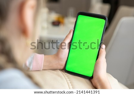 Little girl use smart phone with blank green screen mock up display for advertising text while rest on couch in living room at modern home. Phone with green screen and chroma key for copy space