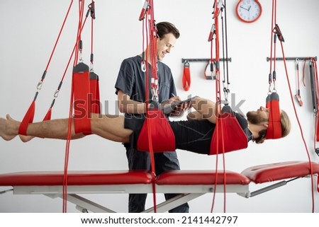Rehabilitation specialist examining male patient before active treatment on suspension straps. Therapeutic exercises and neuromuscular activation on red cord slings Royalty-Free Stock Photo #2141442797