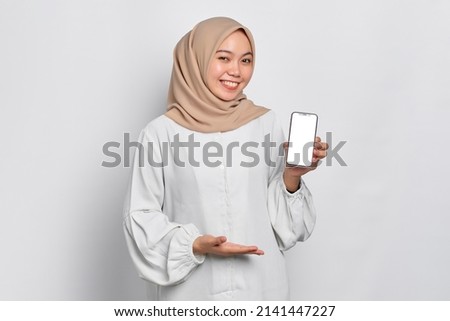 Smiling young Asian Muslim woman showing mobile phone blank screen recommending App isolated over white background