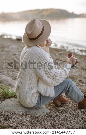 A traveler dressed in cowboy style sitting on the shore and taking a pictures of a sunset on her film camera.