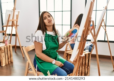 Young hispanic artist women painting on canvas at art studio looking sleepy and tired, exhausted for fatigue and hangover, lazy eyes in the morning. 