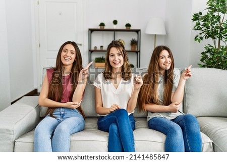 Group of three hispanic girls sitting on the sofa at home with a big smile on face, pointing with hand and finger to the side looking at the camera. 