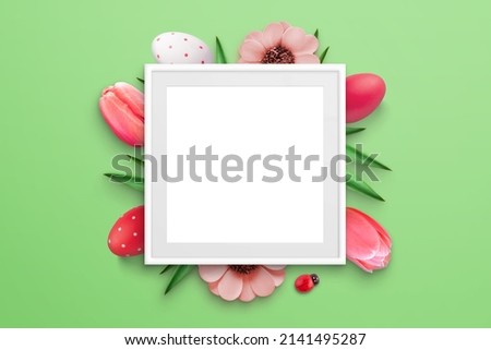 Blank picture frame surrounded by Easter eggs and flowers on green surface. Top view, flat lay composition