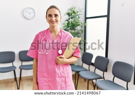 Young nurse woman at medical clinic waiting room looking positive and happy standing and smiling with a confident smile showing teeth 