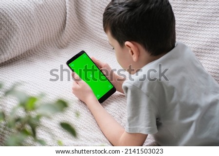 The boy looks at the screen of the phone with a chrome key indoors. An unrecognizable child uses a smartphone with a green screen to watch a video.