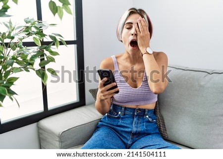 Young beautiful woman using smartphone typing message sitting on the sofa yawning tired covering half face, eye and mouth with hand. face hurts in pain. 
