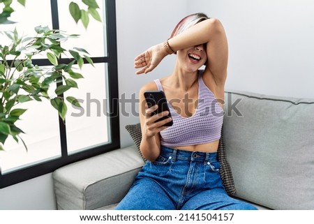 Young beautiful woman using smartphone typing message sitting on the sofa smiling cheerful playing peek a boo with hands showing face. surprised and exited 